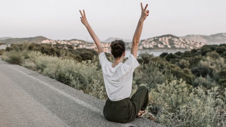 Woman on a roadside in Antalya raises peace sign, embracing summer with mountain views and tranquility.